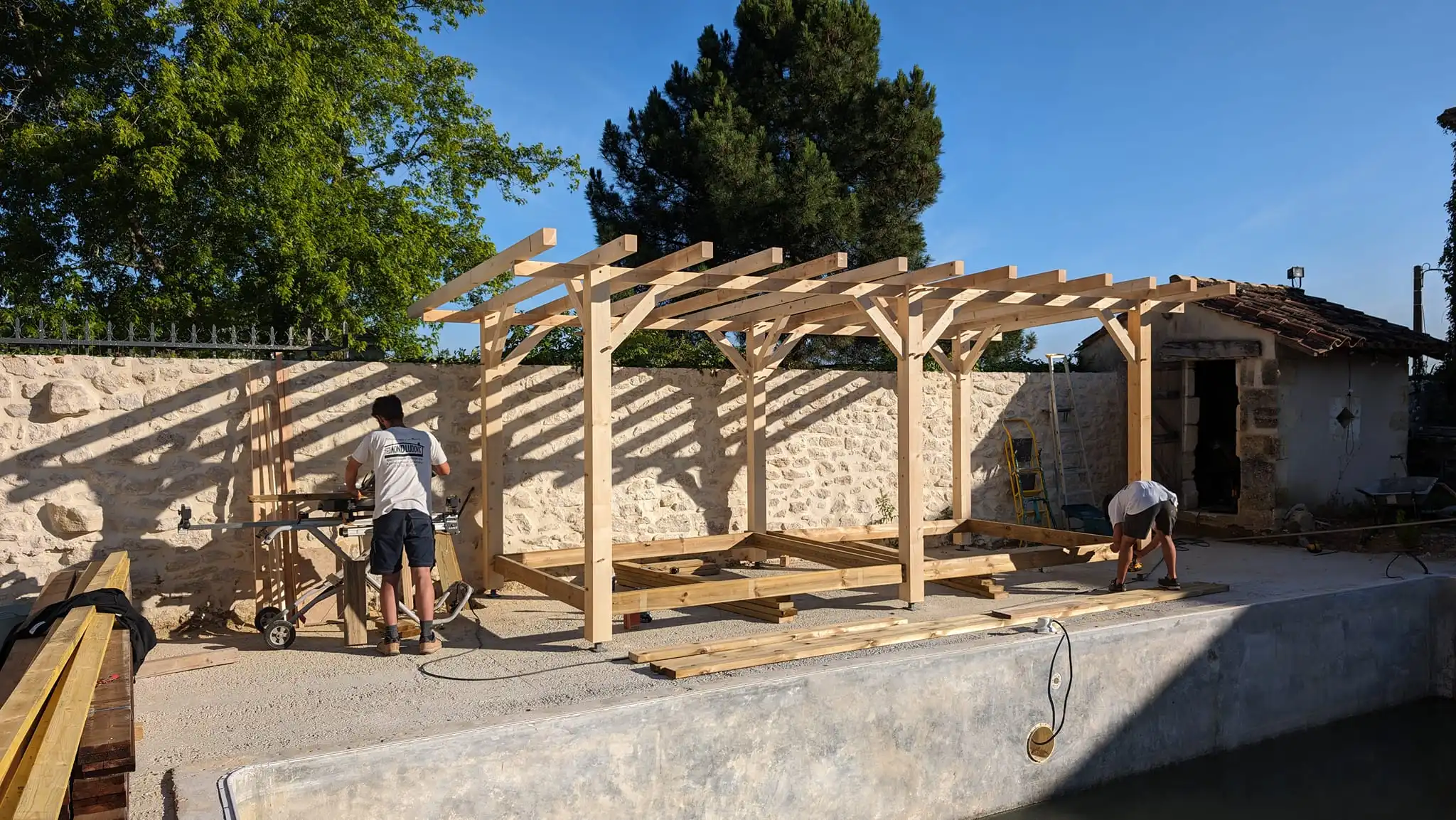 Deux ouvriers construisent une pergola en bois clair près d'un mur en pierre et d'une petite bâtisse ancienne, sous un ciel bleu.