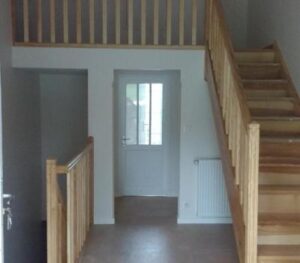 Intérieur d'une maison neuve avec escalier en bois clair et garde-corps assorti menant à une mezzanine. Porte vitrée blanche au centre et radiateur.