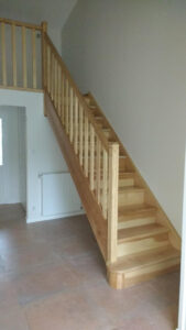 Escalier droit en bois clair avec rambarde à balustres verticaux, menant à une mezzanine. Plancher de carreaux terre cuite et radiateur blanc.