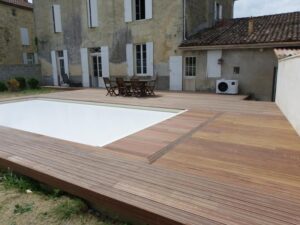 Terrasse en bois exotique neuf autour d'une piscine couverte par une bâche blanche, devant une vieille maison aux volets blancs.