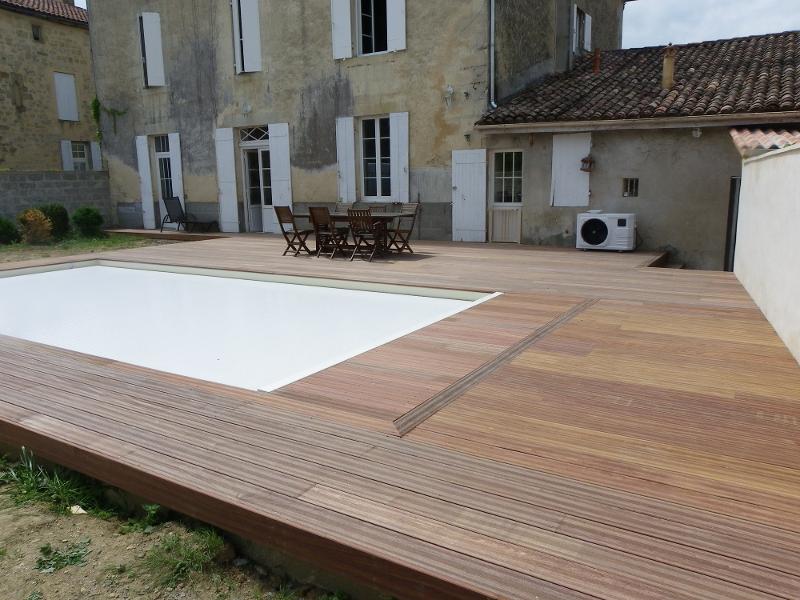 Terrasse en bois exotique neuf autour d'une piscine couverte par une bâche blanche, devant une vieille maison aux volets blancs.