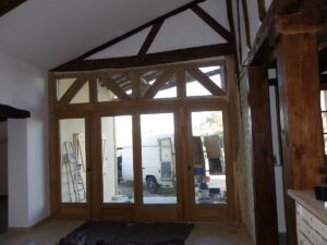 Intérieur en rénovation avec grande baie vitrée à croisillons en bois clair, poteaux de charpente sombres et escalier visible à travers le verre.