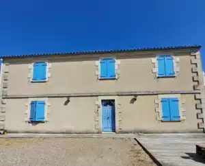 Façade d'une maison traditionnelle beige sous un ciel bleu vif, avec des volets bleu turquoise, une porte bleue assortie et des chaînes d'angle en pierre.