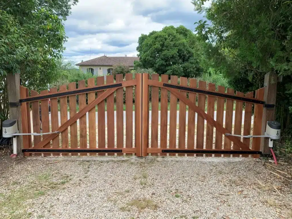 Portail battant automatique en bois pour entrée Portail battant en bois brun avec automatisation, ouverture vers une maison à l'arrière-plan sous un ciel nuageux.