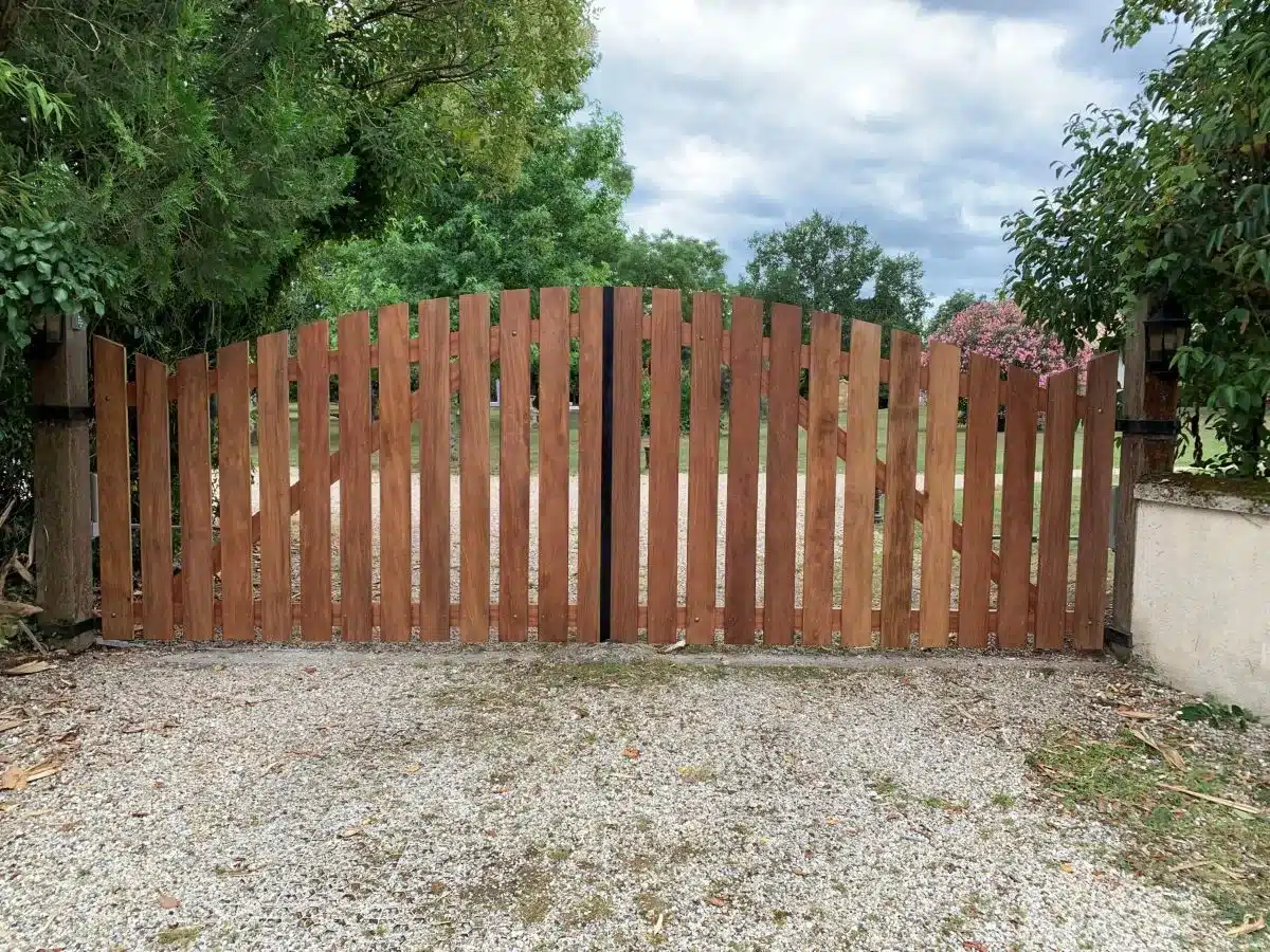Portail double en bois brun clair avec planches verticales et sommet arqué, ouvrant sur une allée de gravier, encadré par des arbres verts et un ciel nuageux.