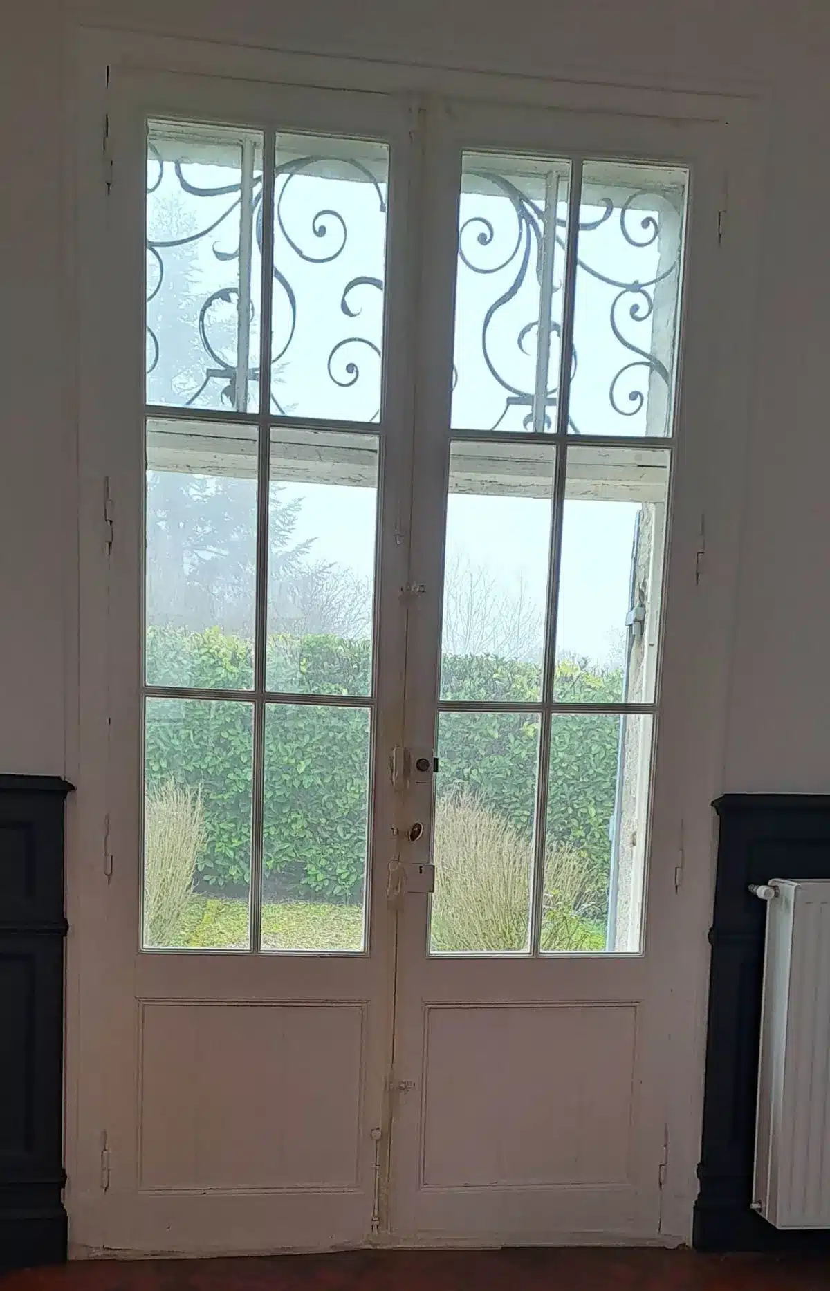 Double porte-fenêtre vitrée blanche à meneaux, avec grille décorative en fer forgé en partie haute, donnant sur un jardin brumeux avec haie verte.