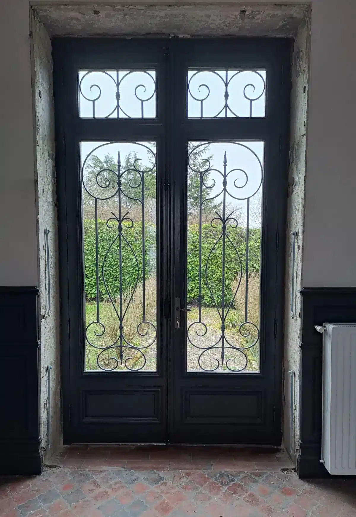Double porte vitrée noire de style ancien avec barreaux en fer forgé ornés de volutes, donnant sur un jardin verdoyant, devant un sol en carreaux de terre cuite.