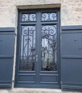 Double porte vitrée grise avec ferronnerie décorative de style fleur de lys, encadrée par des volets en bois bleu-gris et une maçonnerie en pierre.